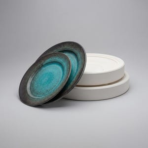 May include: Two oval ceramic plates with a turquoise center and dark gray rim are stacked in front of a stack of white ceramic serving dishes. The plates have a glossy finish. The serving dishes are round with a flat top.