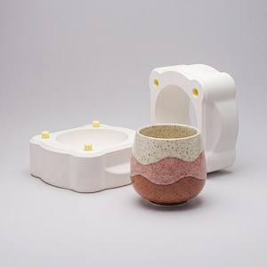 May include: A ceramic cup with a speckled cream, pink, and terracotta wavy pattern sits in front of a white plaster mold. The cup has a rounded shape and a smooth finish, while the mold features yellow alignment pins for casting pottery.
