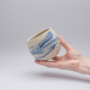 Könnte beinhalten: Eine kleine, abgerundete Keramiktasse mit einer gesprenkelten cremefarbenen Basis und einem wirbelnden blauen Glasurmuster. Die Tasse hat eine breite, offene Oberseite und eine glatte, geschwungene Außenfläche mit sichtbaren dunklen Sprenkeln.