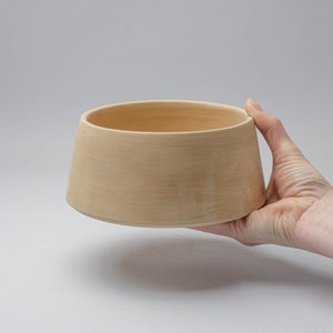 Puede incluir: Un cuenco de cerámica marrón claro con una base ancha y redondeada y un borde ligeramente acampanado. El cuenco se sostiene en una mano, mostrando su tamaño y forma.