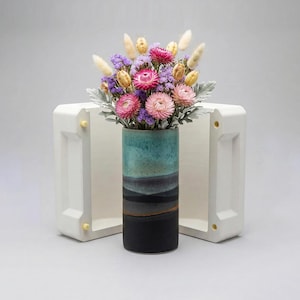 Puede incluir: Un jarrón cilíndrico de cerámica, con una combinación de colores turquesa, gris y negro, contiene un ramo de flores secas. El arreglo presenta flores rosas, moradas y amarillas, con acentos blancos plumosos. El jarrón está sobre un fondo blanco geométrico.