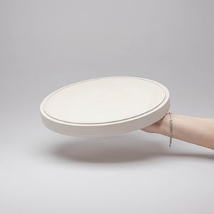May include: A circular, off-white tray with a smooth, matte finish and a raised outer rim. The tray features a flat, recessed centre surface and a thick, solid edge, presenting a clean, neutral appearance suitable for various display purposes.