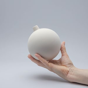 Puede incluir: Un jarrón de cerámica blanca con forma de esfera y una pequeña abertura en la parte superior.