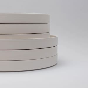 May include: A stack of five circular, off-white ceramic plates with smooth, flat rims and a matte finish. The plates are layered vertically, showcasing their uniform thickness and clean, rounded edges in a simple, stacked arrangement.