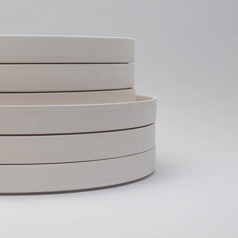 May include: A stack of five circular, off-white ceramic plates with smooth, flat rims and a matte finish. The plates are layered vertically, showcasing their uniform thickness and clean, rounded edges in a simple, stacked arrangement.