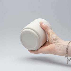 Op de afbeelding: Een witte, cilindrische keramische container met een afgeronde basis. De container wordt in een hand gehouden, waarbij de gladde, matte afwerking wordt getoond. Het ontwerp is eenvoudig en modern, geschikt voor diverse toepassingen.