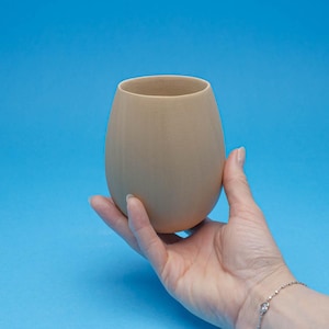 Può includere: Una tazza in ceramica di colore marrone chiaro con una forma arrotondata, simile a un uovo, tenuta nella mano di una persona.