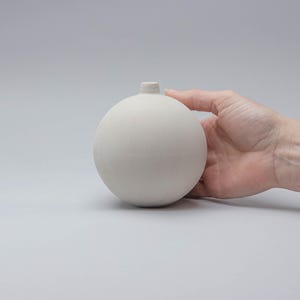 Puede incluir: Un pequeño jarrón esférico de cerámica blanquecina con un cuello corto y cilíndrico. El jarrón está sostenido por una mano, mostrando su acabado liso y mate. El diseño minimalista sugiere una estética moderna, adecuada para pequeños arreglos florales o exhibición decorativa.