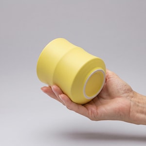 Puede incluir: Una taza de cerámica de color amarillo mate con una forma cilíndrica lisa y ligeramente cónica. La base de la taza presenta un borde circular blanco limpio que contrasta con el exterior amarillo sólido. El diseño es sencillo, funcional y uniforme.