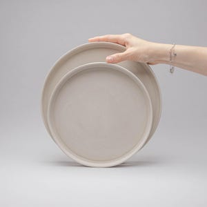 May include: Two circular, light beige ceramic plates with a smooth, matte finish and raised edges. The plates are stacked slightly offset, featuring a simple, clean design with a uniform colour throughout the surface of the dinnerware set.