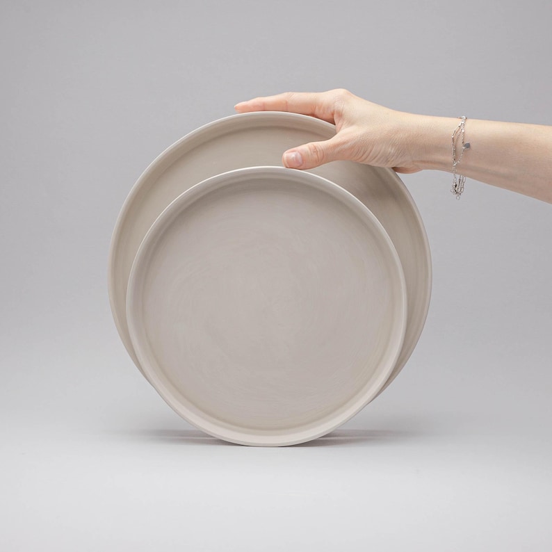 May include: Two circular, light beige ceramic plates with a smooth, matte finish and raised edges. The plates are stacked slightly offset, featuring a simple, clean design with a uniform colour throughout the surface of the dinnerware set.