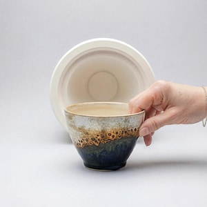 May include: A handcrafted ceramic bowl with a distinctive glaze. The bowl displays a colour gradient of cream, brown, and dark blue. A hand holds the bowl, with a larger, similar bowl visible in the background. The bowl is likely for serving food or as a decorative piece.