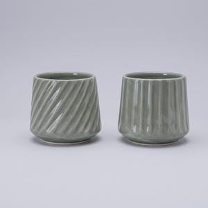 May include: Two sage green ceramic cups with a glossy finish. The cup on the left features a diagonal, spiraling ribbed texture, while the cup on the right displays vertical, straight fluted ridges. Both vessels have a wide, open top.