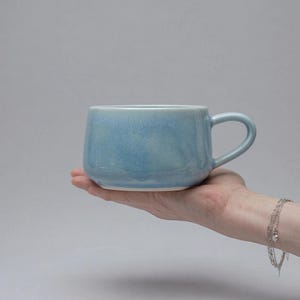 Puede incluir: Una taza de cerámica azul claro con forma redondeada y asa. La taza se sostiene en una mano sobre un fondo gris. La taza tiene un acabado liso y brillante y un interior blanco.