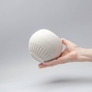 May include: A white, egg-shaped ceramic bowl with vertical grooves near the base. The bowl is held in a hand against a light gray background. The bowl's smooth surface contrasts with the textured grooves, creating a minimalist aesthetic.