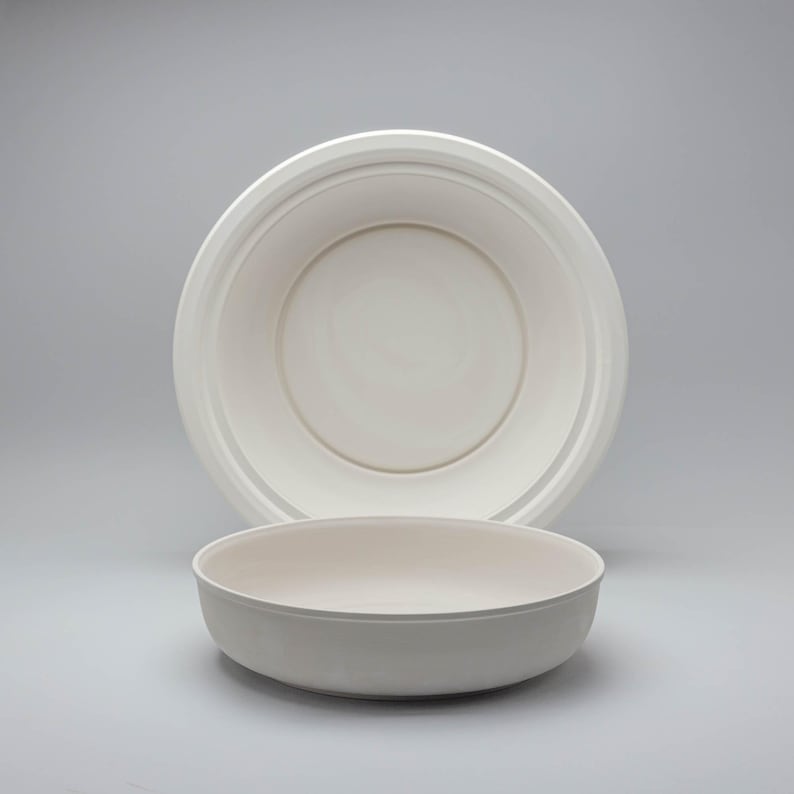 Pode incluir: Duas tigelas redondas brancas s&atilde;o exibidas em um fundo cinza claro. Uma tigela est&aacute; em primeiro plano e a outra atr&aacute;s. As tigelas t&ecirc;m um design simples e limpo, adequado para servir alimentos.