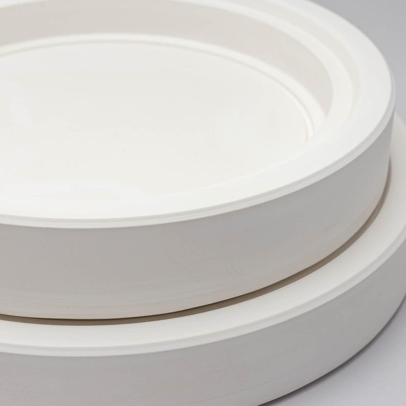 May include: A stack of two circular white ceramic plates with smooth, matte surfaces. Each plate features a wide, flat rim and a recessed centre section, creating a clean, uniform appearance with no additional patterns or decorative elements.