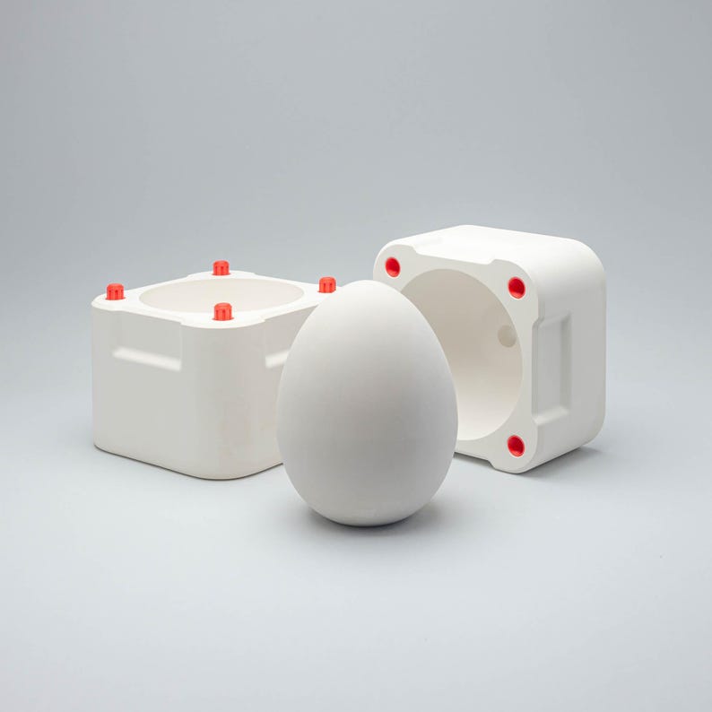 May include: A white ceramic egg mold with a removable base. The mold has five red plastic knobs on the top and bottom. The egg mold is sitting on a white surface.