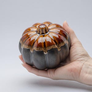 Może przedstawiać: Ceramiczny pojemnik w kształcie dyni z błyszczącym, dwutonowym wykończeniem. Wierzch jest bogaty brązowy z jaśniejszymi akcentami, a podstawa ciemnoszara. Dekoracyjny przedmiot na jesień.