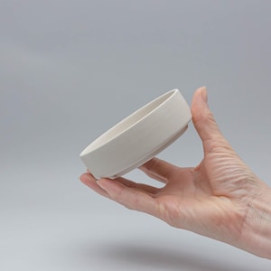 Könnte beinhalten: Eine kleine, cremefarbene Keramikschale mit einem breiten, flachen Design. Die Schale wird in einer Hand gehalten und zeigt ihre schlichte, moderne Ästhetik. Der Rand der Schale ist leicht abgewinkelt, und die Oberfläche hat eine subtile Textur.