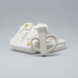 May include: A white ceramic pitcher with a handle and a heart-shaped design, next to a white mold with yellow accents. The pitcher is centered, with the mold in the background. The background is a solid light gray.