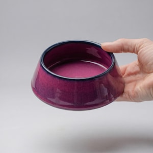 May include: A handmade, ceramic bowl with a glossy, gradient finish in shades of magenta and purple. The bowl has a dark blue rim and a unique, angled shape. The bowl is held by a hand against a neutral background.