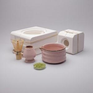 Könnte beinhalten: Eine hellrosa Matcha-Schale aus Keramik mit Ausguss, ein passender rosa Besenhalter, ein Matcha-Besen aus Bambus und eine kleine durchsichtige Schale mit grünem Matcha-Pulver. Dahinter befinden sich zwei weiße Schutzverpackungsblöcke aus Schaumstoff.