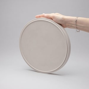 May include: A circular, light beige tray with a smooth, matte finish and a raised outer rim. The tray features a flat, solid surface and a consistent, neutral colour throughout, presenting a simple, functional design for holding various small items.