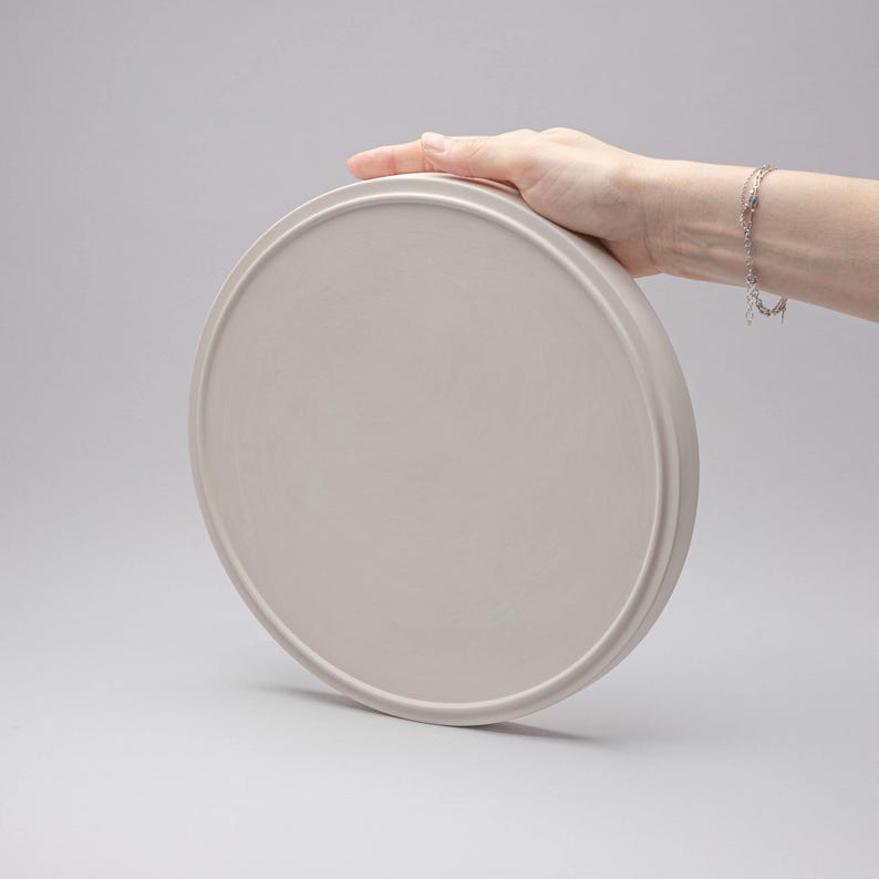 May include: A circular, light beige tray with a smooth, matte finish and a raised outer rim. The tray features a flat, solid surface and a consistent, neutral colour throughout, presenting a simple, functional design for holding various small items.
