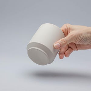 Op de afbeelding: Een lichtbeige keramische mok met een cilindrisch lichaam en een licht uitlopende basis. De mok wordt door een hand vastgehouden, waarbij de gladde, matte afwerking wordt getoond. Het ontwerp is minimalistisch en modern, geschikt voor dranken.