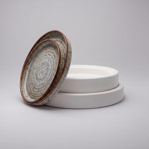 May include: Two circular ceramic plates with a speckled grey and white finish and a dark brown rim, positioned upright against a stack of two plain white, smooth-surfaced ceramic plates. The plates feature a subtle concentric ring texture.