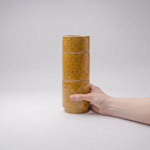 May include: A stack of three cylindrical ceramic cups in a mustard yellow glaze with dark brown speckles. Each cup features a smooth, straight-sided design with a slightly recessed base, allowing them to fit securely together in a vertical column.