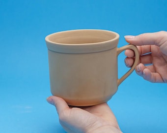 Puede incluir: Una taza de cerámica de color marrón claro sin decoración, con asa. La taza está siendo sostenida en la mano de una persona.