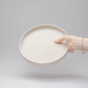 May include: A circular, off-white tray with a smooth, matte finish and a raised edge. The tray features a flat, wide base and a consistent, neutral colour throughout, presenting a simple, functional design for holding various small items.