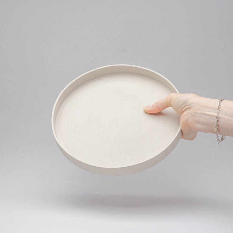 May include: A circular, off-white tray with a smooth, matte finish and a raised edge. The tray features a flat, wide base and a consistent, neutral colour throughout, presenting a simple, functional design for holding various small items.