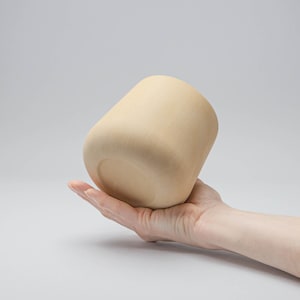 Puede incluir: Una maceta de cerámica beige, cilíndrica, con base redondeada. La maceta se sostiene en una mano, mostrando su superficie lisa y sin esmaltar. El diseño de la maceta es simple y moderno, adecuado para plantas o uso decorativo.