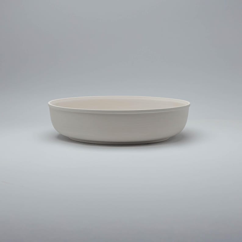 Pode incluir: Uma tigela de cer&acirc;mica branca suja. A tigela tem uma forma rasa e arredondada com um acabamento liso e fosco. A tigela &eacute; adequada para servir alimentos ou como pe&ccedil;a decorativa. A cor neutra da tigela torna-a vers&aacute;til.