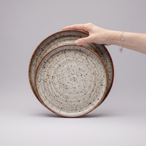 May include: Two round ceramic plates with a speckled cream finish and dark brown flecks. The plates feature a concentric ridged texture on the surface and a dark brown rim. The design is consistent across both plates, which are stacked together.