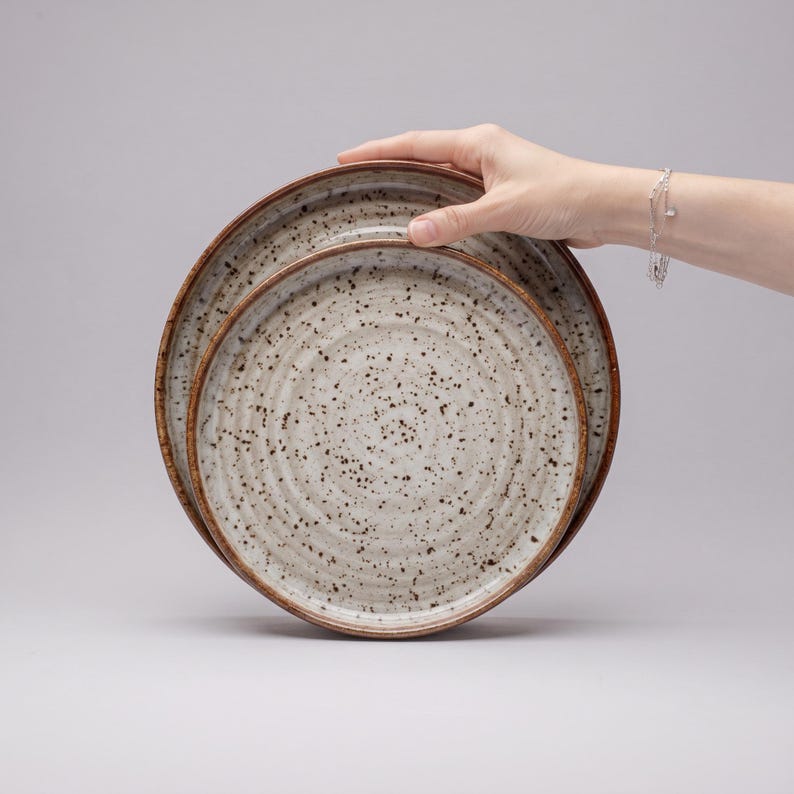 May include: Two round ceramic plates with a speckled cream finish and dark brown flecks. The plates feature a concentric ridged texture on the surface and a dark brown rim. The design is consistent across both plates, which are stacked together.