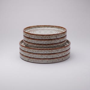 May include: A stack of six round ceramic plates featuring a speckled off-white surface with dark brown flecks and a distinct brown rim. The plates are arranged in two groups, with three smaller plates stacked on top of three larger plates.