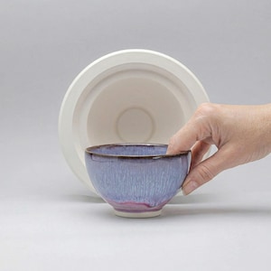 Pode incluir: Uma pequena tigela de cerâmica feita à mão com um esmalte azul claro e uma borda marrom escura. A tigela tem uma base rosa-púrpura e é segurada perto de uma peça de cerâmica branca maior. A tigela tem cerca de 7,6 cm de diâmetro.