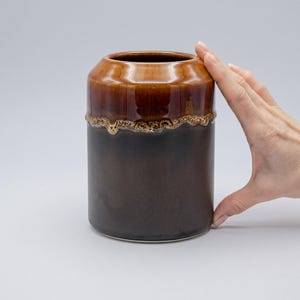 Puede incluir: Un jarrón cilíndrico de cerámica con base marrón oscuro y parte superior marrón claro. La parte superior tiene una banda texturizada de color dorado. El jarrón mide unos 15 cm de alto.
