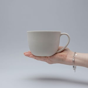 Puede incluir: Una taza grande de cerámica blanquecina con un cuerpo redondeado y un asa curva. La taza se sostiene en la mano abierta de una persona sobre un fondo gris claro. La superficie lisa de la taza contrasta con la textura de la mano. Se ve una pulsera plateada.