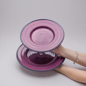May include: Two round ceramic plates featuring a vibrant magenta and purple swirled pattern with a dark navy blue rim. The plates have a glossy finish and a slightly raised edge, showcasing a hand-painted aesthetic with visible circular brush strokes.
