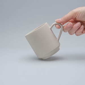 以下が含まれることがあります： 丸みを帯びた取っ手が付いた白いセラミック製のマグカップ。マグカップは少し傾いており、質感のある素材でできているように見える。