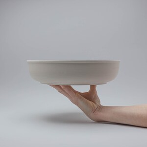 Pode incluir: Uma tigela de cer&acirc;mica cinza claro com um design largo e raso. A tigela &eacute; segurada em uma m&atilde;o contra um fundo cinza liso. O di&acirc;metro da tigela &eacute; de aproximadamente 30 cm.