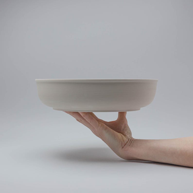 Pode incluir: Uma tigela de cer&acirc;mica cinza claro com um design largo e raso. A tigela &eacute; segurada em uma m&atilde;o contra um fundo cinza liso. O di&acirc;metro da tigela &eacute; de aproximadamente 30 cm.