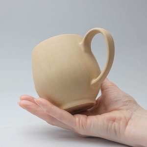 May include: A light beige ceramic mug with a rounded body and a slightly curved handle.