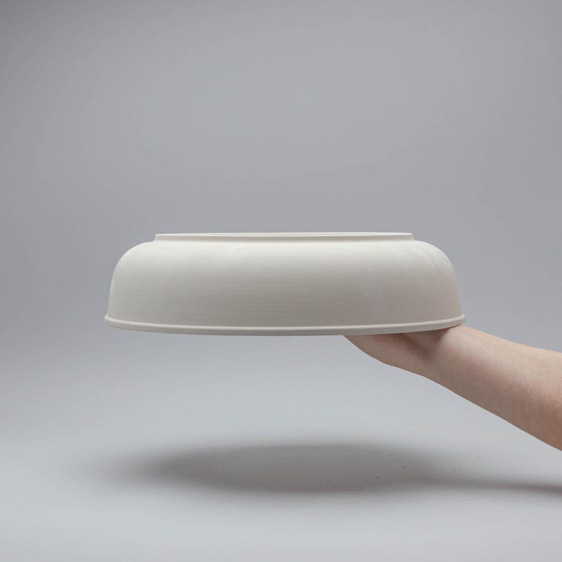 Pode incluir: Uma tigela de cer&acirc;mica branca com base arredondada e borda plana. A tigela &eacute; segurada por uma m&atilde;o contra um fundo cinza. O di&acirc;metro da tigela &eacute; de aproximadamente 30 cm.