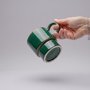 Può includere: Una tazza in ceramica verde smeraldo profondo e lucido, caratterizzata da un corpo cilindrico con un distinto rilievo orizzontale vicino alla base. La tazza presenta un manico curvo e una finitura liscia e riflettente con smalto scuro.